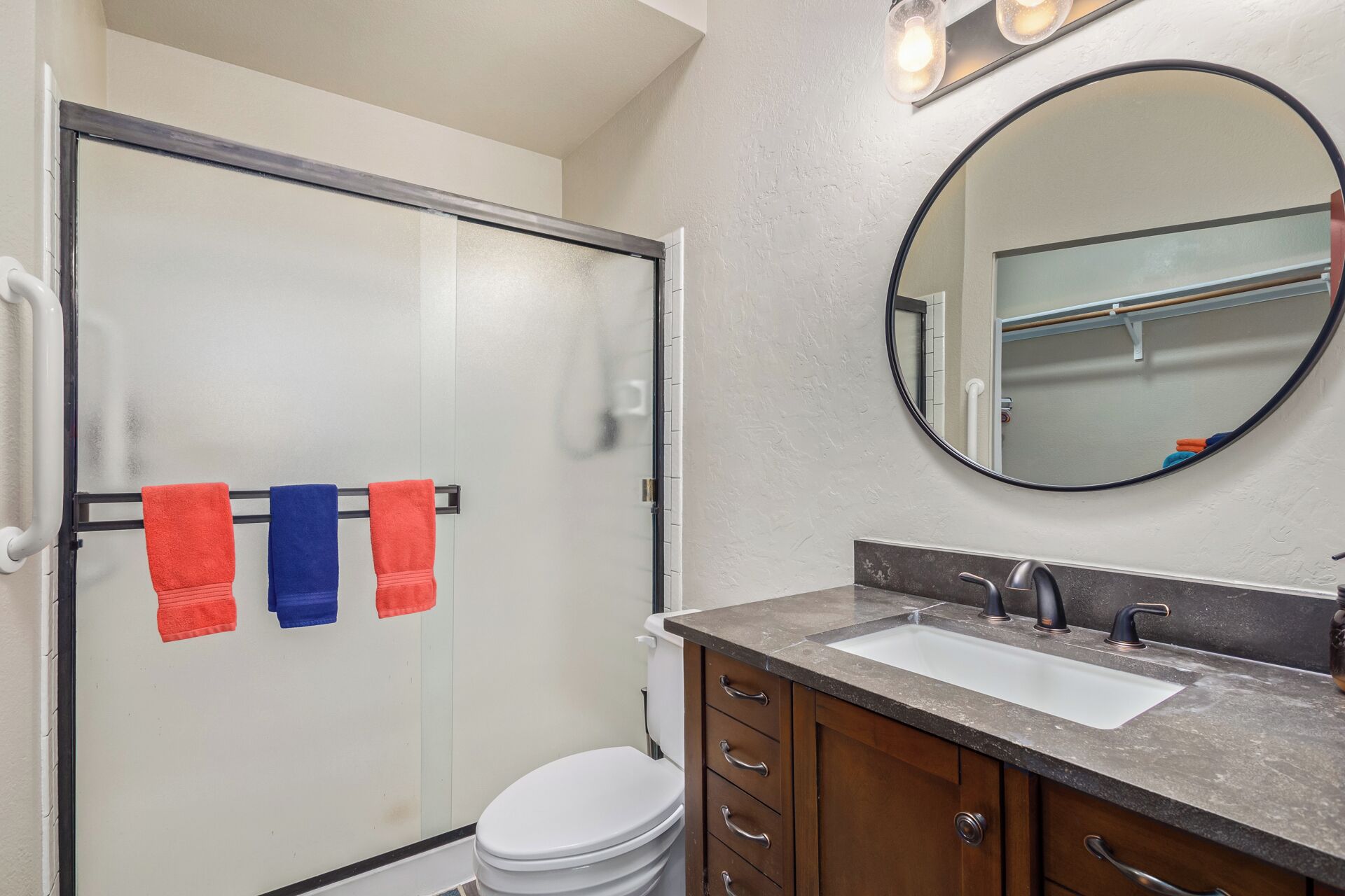 Une salle de bain avec une porte de douche en verre, des toilettes blanches, un lavabo avec un plan de toilette foncé et un miroir rond.