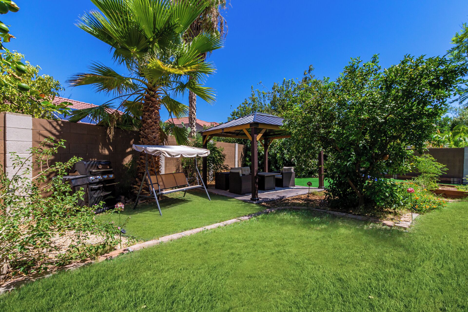 A backyard with a grill, a swing, a covered seating area, and a palm tree.