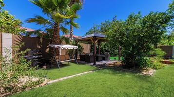 A backyard with a grill, a swing, a covered seating area, and a palm tree.