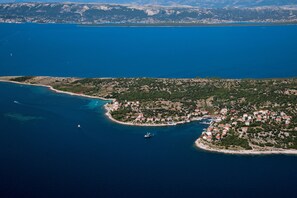 An aerial view of a coastal town with a marina, surrounded by clear blue waters and green hills in the background. - Apartments Mario (Novalja)