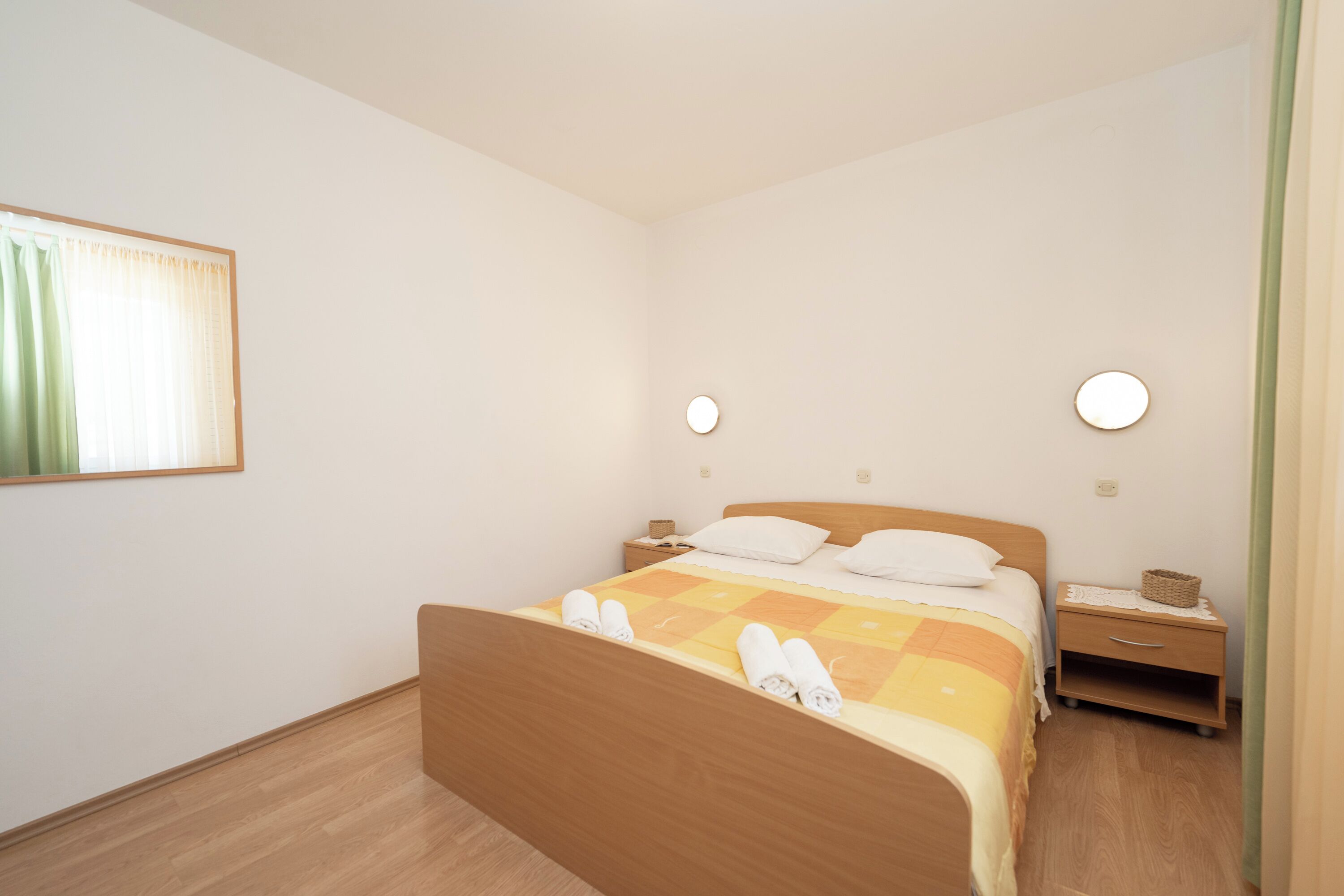 A bedroom with a wooden bed, a nightstand, a wall-mounted light, and a window with curtains.