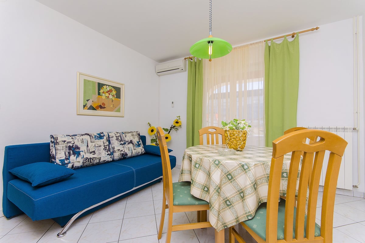 A room with a blue sofa, a wooden dining table with a checkered tablecloth, and a green hanging light fixture.