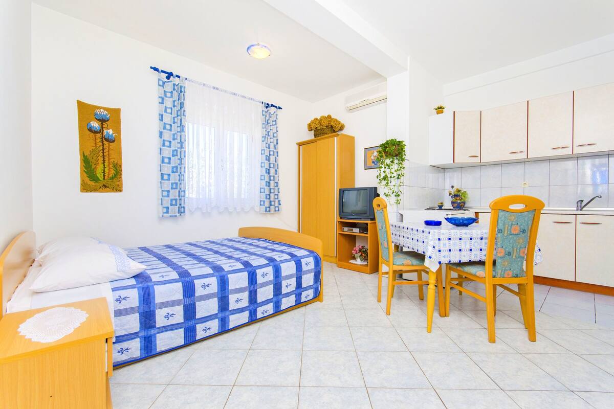A compact room with a bed, a small table with chairs, a television, and a kitchenette.