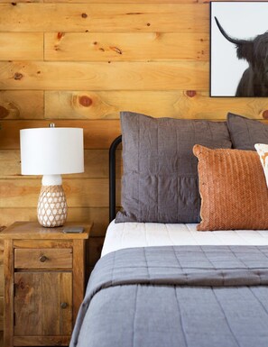 Una habitación con paneles de madera, una cama, almohadas, una mesita de noche con una lámpara y un cuadro en la pared.