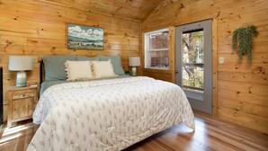 Un dormitorio de cabaña de madera con una cama, dos lámparas de noche, un ventanal con persianas y un cuadro en la pared.