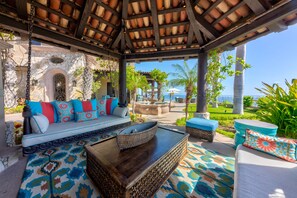 Ein überdachter Außenbereich mit einer Steinsäule, einer Couch mit bunten Kissen, einem gewebten Couchtisch und Blick auf das Meer.