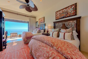 Een hotelkamer met een groot bed, een balkon met uitzicht op zee, een plafondventilator en kunstwerken aan de wanden.