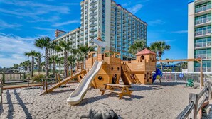 A beach playground with a slide and climbing structures, surrounded by palm trees and a tall hotel building in the background. - 🐠Oceanfront | Indoor Heated Water Park + Lazy River | Dunes Village Resort 701 (Myrtle Beach)
