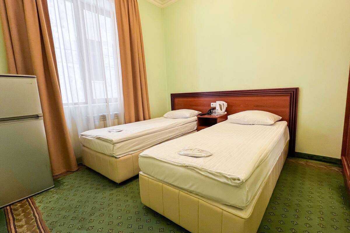 A hotel room with two single beds, a small bedside table with a lamp, a refrigerator, and a window with curtains.