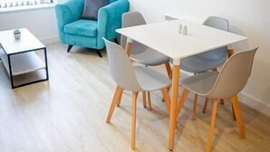 A modern dining area with a white table and grey chairs, a blue sofa, and a painting on the wall. - One Bed Serviced Apartment Preston (Preston)