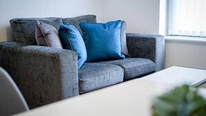 A grey sofa with blue and grey cushions, a white table, and a window with blinds. - One Bed Serviced Apartment Preston (Preston)