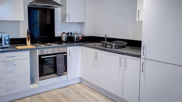 A modern kitchen with white cabinets, a stainless steel range hood, a built-in oven, and a black countertop. - One Bed Serviced Apartment Preston (Preston)