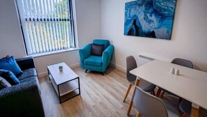 A modern living room with a sofa, a teal armchair, a coffee table, and a dining area with a table and chairs. - One Bed Serviced Apartment Preston (Preston)