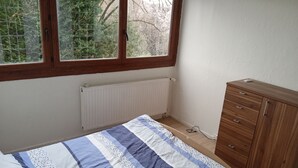 Een slaapkamer met een bed, een houten dressoir, een radiator en een raam met uitzicht op bomen.