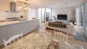 Une cuisine moderne dotée d’un îlot central, un coin repas avec une table et des chaises, un salon avec un canapé et une télévision, et de grandes fenêtres qui laissent entrer la lumière naturelle.