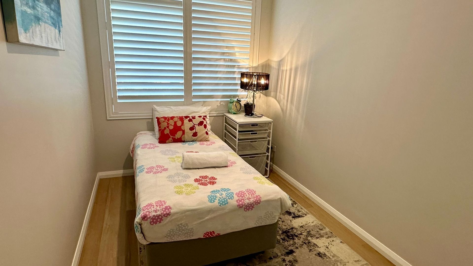 A small bedroom with a bed, a nightstand, a lamp, and a window with blinds.