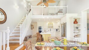 A spacious dining area with a high ceiling fan, a staircase leading to an upper level, and a kitchen in the background.