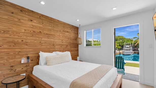 A bedroom with a wooden headboard, a bed with white bedding, a small round table, and a view of a swimming pool through a door.