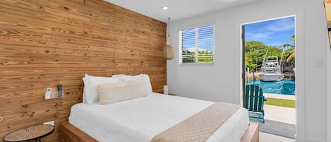 Un dormitorio con cabecera de madera, cama con ropa blanca, una mesita redonda y vista a una piscina a través de una puerta.
