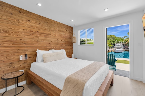 A bedroom with a wooden headboard, a bed with white bedding, a small round table, and a view of a swimming pool through a door.