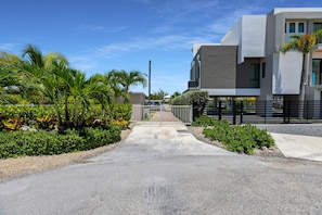 Un quartier résidentiel moderne avec une allée pavée menant à une entrée sécurisée par un portail, le tout entouré d’une végétation luxuriante et de palmiers.