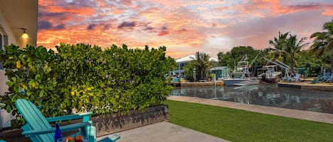 A waterfront patio with a view of the sunset, a pool, and lounge chairs.