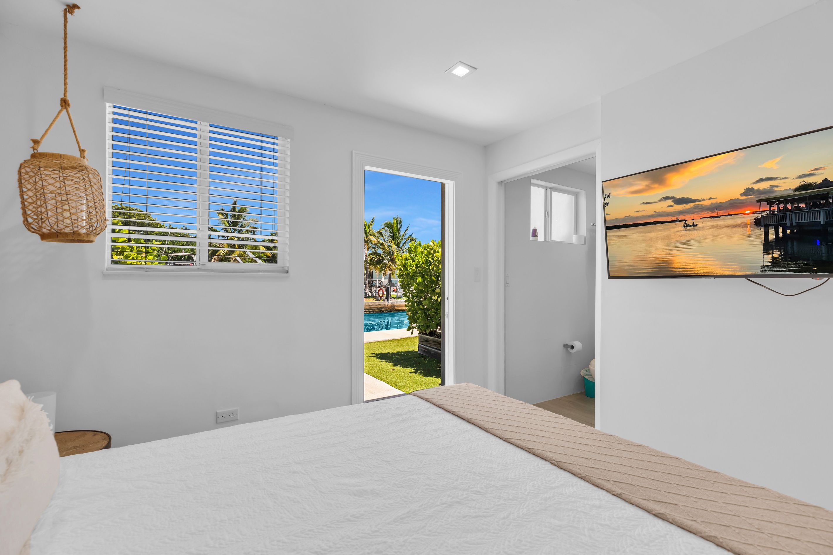 Un dormitorio con cama, ventana con persianas, puerta que da a una piscina y un televisor montado en la pared.