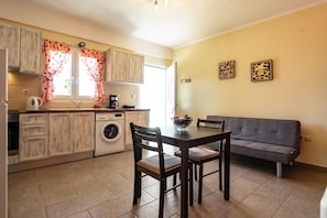 A compact kitchen with wooden cabinets, a washing machine, a dining table with chairs, and a grey sofa.