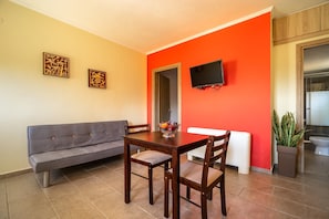 A dining area with a table and chairs, a sofa, a TV mounted on the wall, and a potted plant.