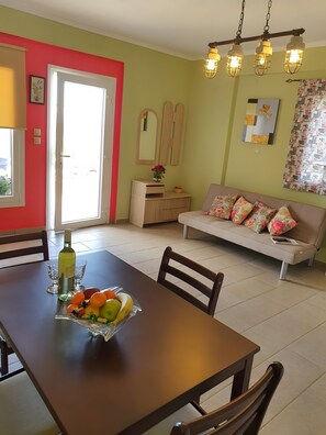 A dining area with a table set for two, a sofa with floral cushions, a mirror, and a hanging light fixture.