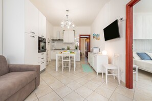 Une cuisine moderne avec des meubles blancs, un coin repas comprenant une table et des chaises, un canapé et un téléviseur fixé au mur.