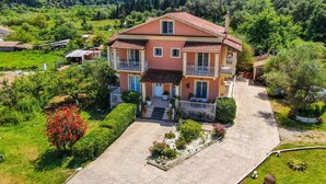 A two-story house with a balcony, surrounded by greenery and a paved driveway. - Apartment "Villa Logas" close to the Beach with Garden & Wi-Fi (Corfu)
