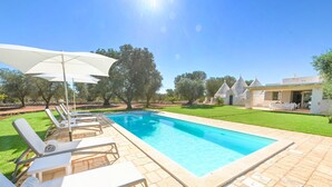 Une piscine entourée de chaises longues et de parasols, elle-même bordée d’une pelouse bien entretenue et de bâtiments en pierre.