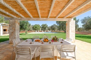 An outdoor dining area with a table set for four, surrounded by white chairs, under a wooden pergola.