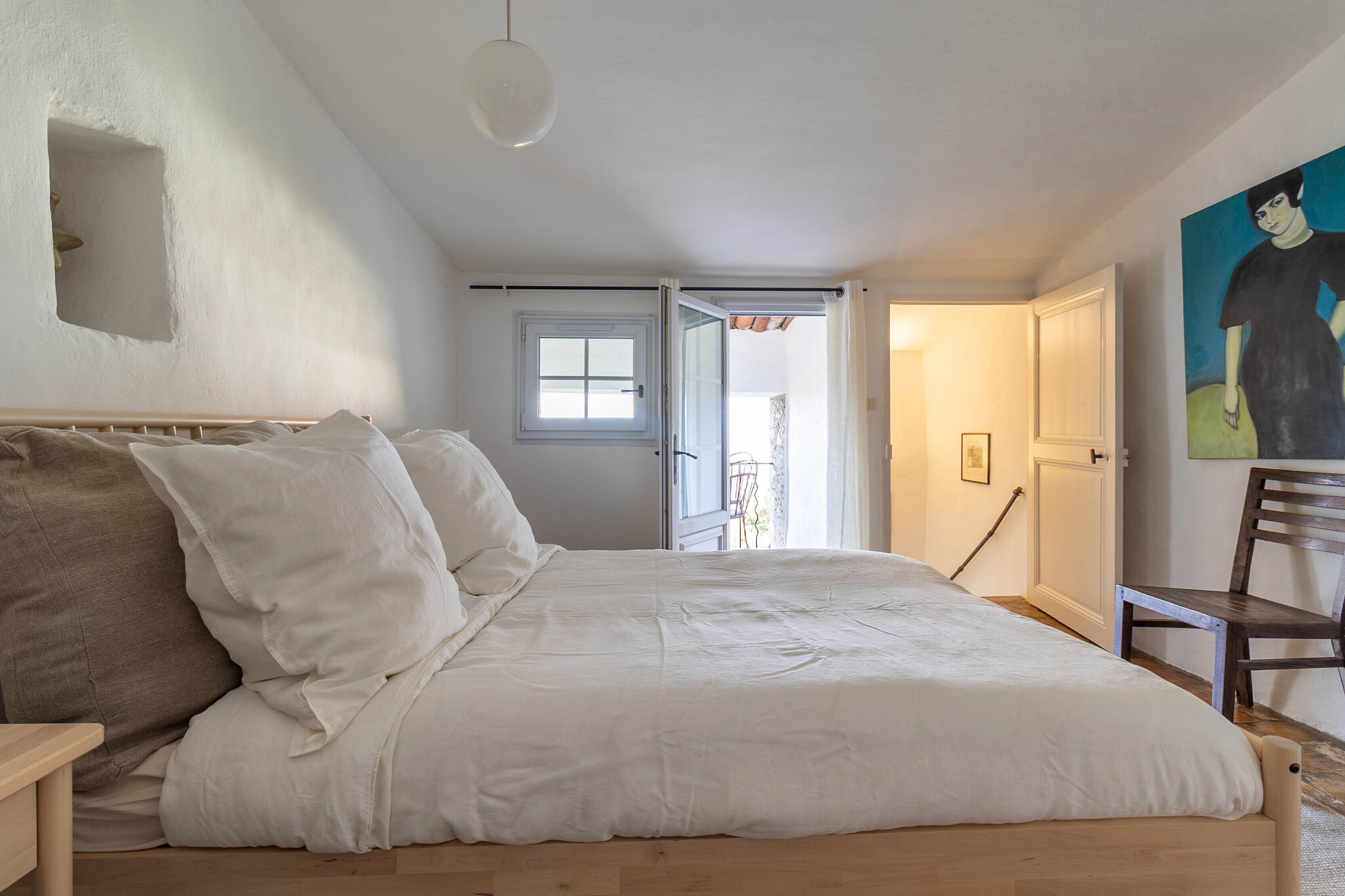 A bedroom with a large bed, a wooden nightstand, a chair, a painting on the wall, and a window with a view.