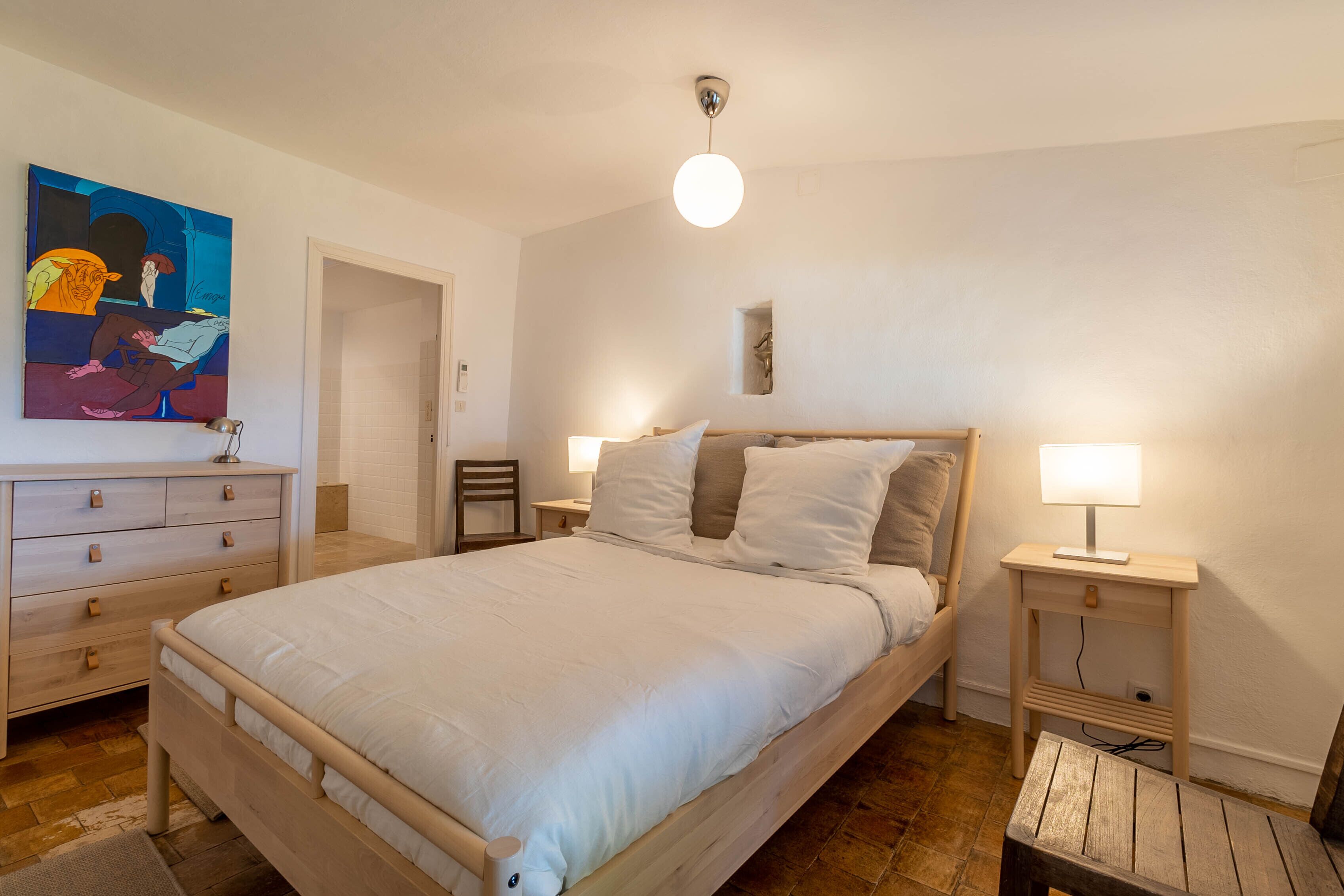 A bedroom with a large bed, two bedside tables with lamps, a wooden dresser, and a painting on the wall.
