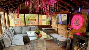 A cozy outdoor seating area with wicker furniture, a glass-top table, and a neon sign. - FINN VILLAGE Mountain View Cottage Garden & XXL HOT TUB (Glasgow)