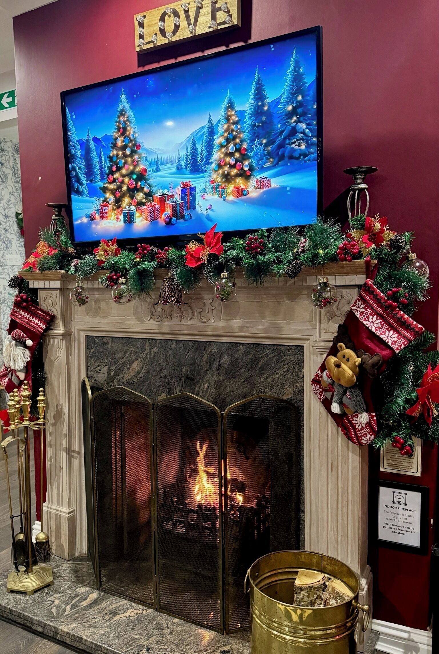 Una chimenea con un televisor encima mostrando una escena navideña, un manto decorado con guirnaldas festivas y calcetines, y un letrero que indica que la chimenea es interior.