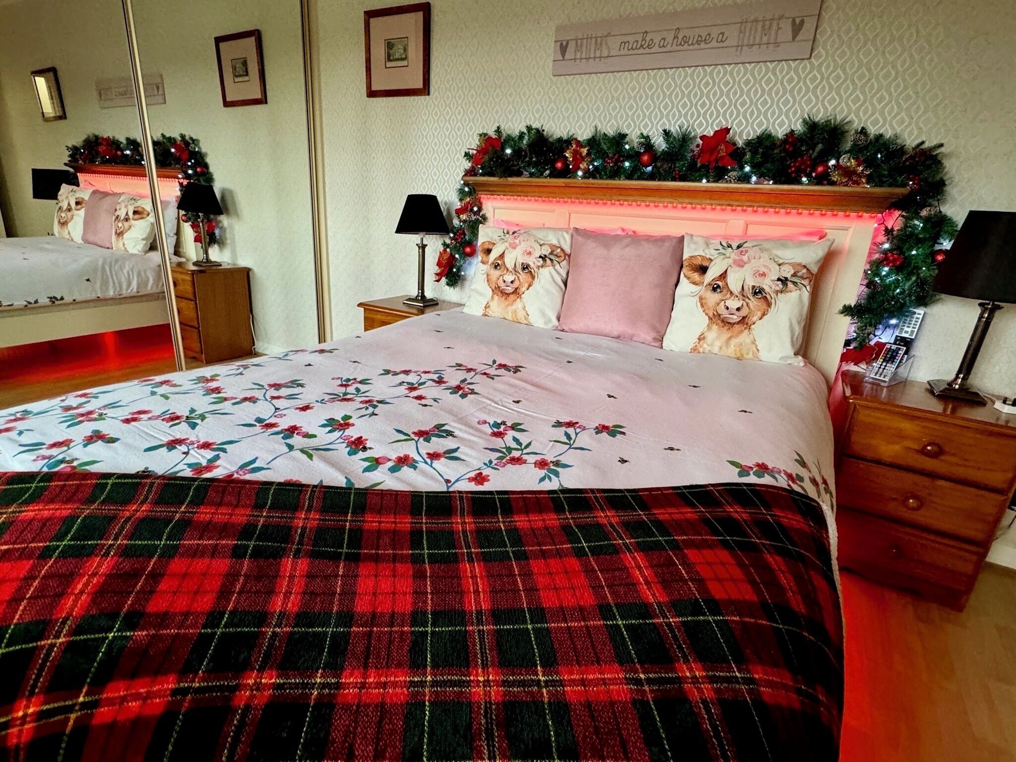 Una cama bien hecha con ropa de cama floral y una manta escocesa, una mesita de noche con lámpara y un espejo en la pared.