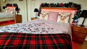 Una cama bien hecha con ropa de cama floral y una manta escocesa, una mesita de noche con lámpara y un espejo en la pared.