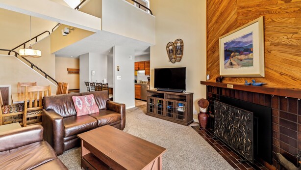 A spacious living room with a brown leather sofa, a wooden coffee table, and a fireplace. A television is mounted on a wooden cabinet, and a staircase leads to an upper level. - Location, Amenities and Value!  Near Ski Area Split Level Condo, Access to Pool, (Steamboat Springs)