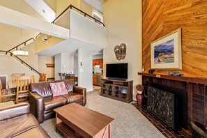 A spacious living room with a brown leather sofa, a wooden coffee table, and a fireplace. A television is mounted on a wooden cabinet, and a staircase leads to an upper level. - Cozy Ski Retreat | Ski Shuttle & Hot Tubs (Steamboat Springs)