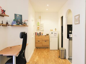 A compact kitchen with a wooden table, a black chair, a white refrigerator, and a trash bin.