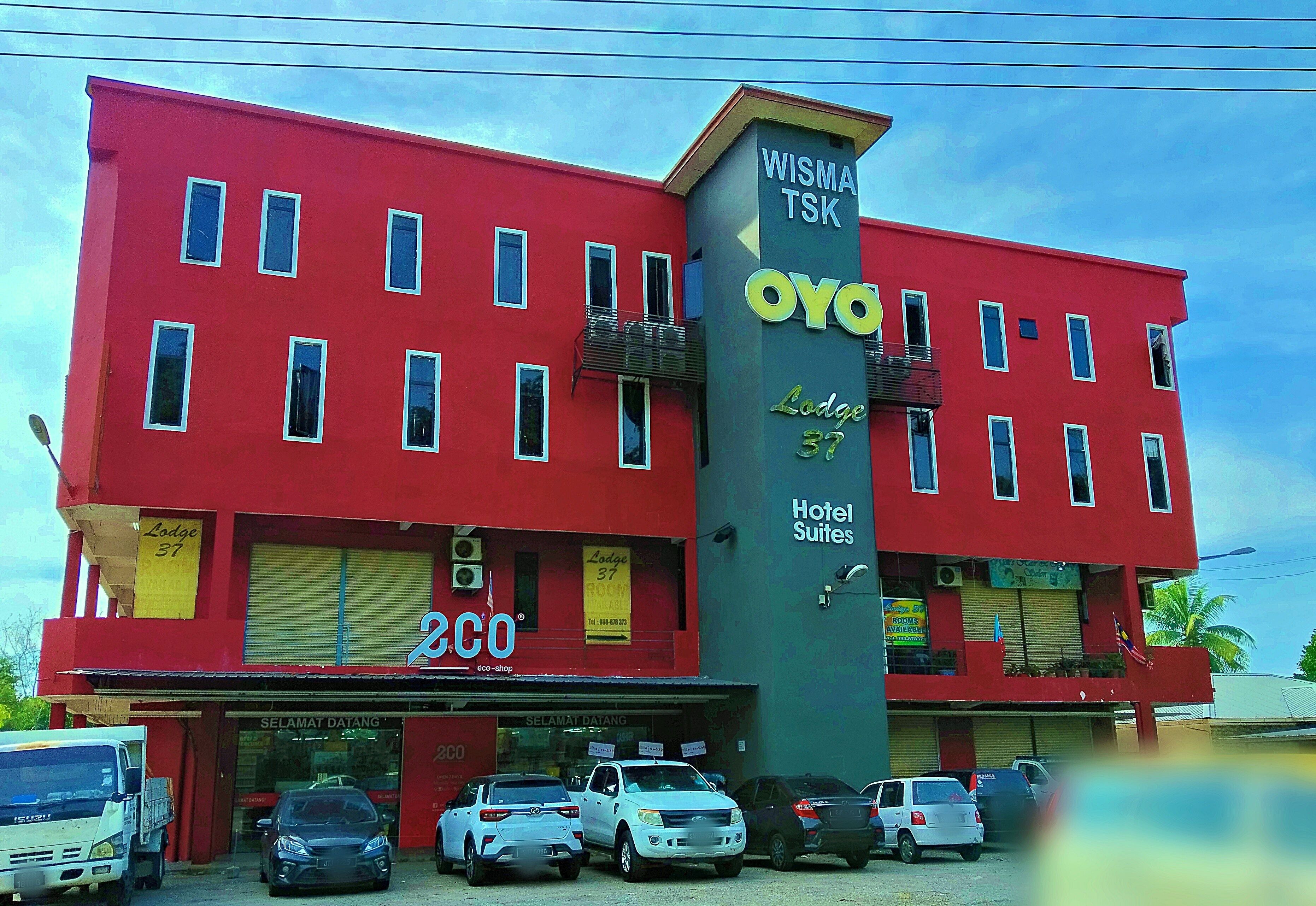Un edificio rojo con múltiples ventanas, un letrero que dice 'WISMA TSK OYO Lodge 37 Hotel Suites' y varios coches estacionados al frente.