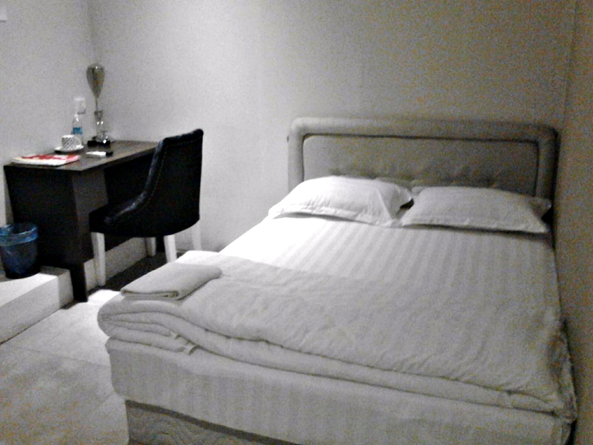 Una cama pequeña y bien hecha con sábanas blancas, un escritorio con una silla y una botella de agua sobre el escritorio.