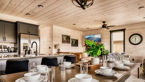 Un comedor amplio con una mesa de madera larga dispuesta para una comida, una cocina con electrodomésticos modernos y un reloj grande en la pared.