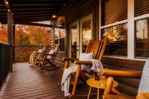 A wooden deck with rocking chairs and a table, overlooking a forest with autumn foliage.