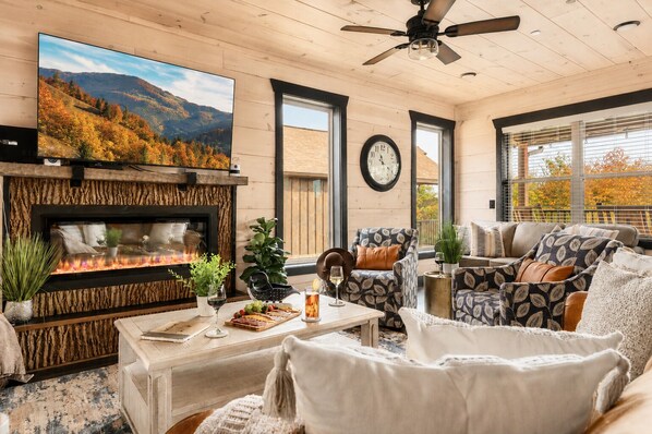 A cozy living room with a fireplace, comfortable seating, a large TV, and a clock on the wall.