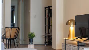 Una sala de estar moderna con una mesa de centro de vidrio, una planta en maceta, un sofá, un televisor sobre un pedestal de mármol y una lámpara dorada.