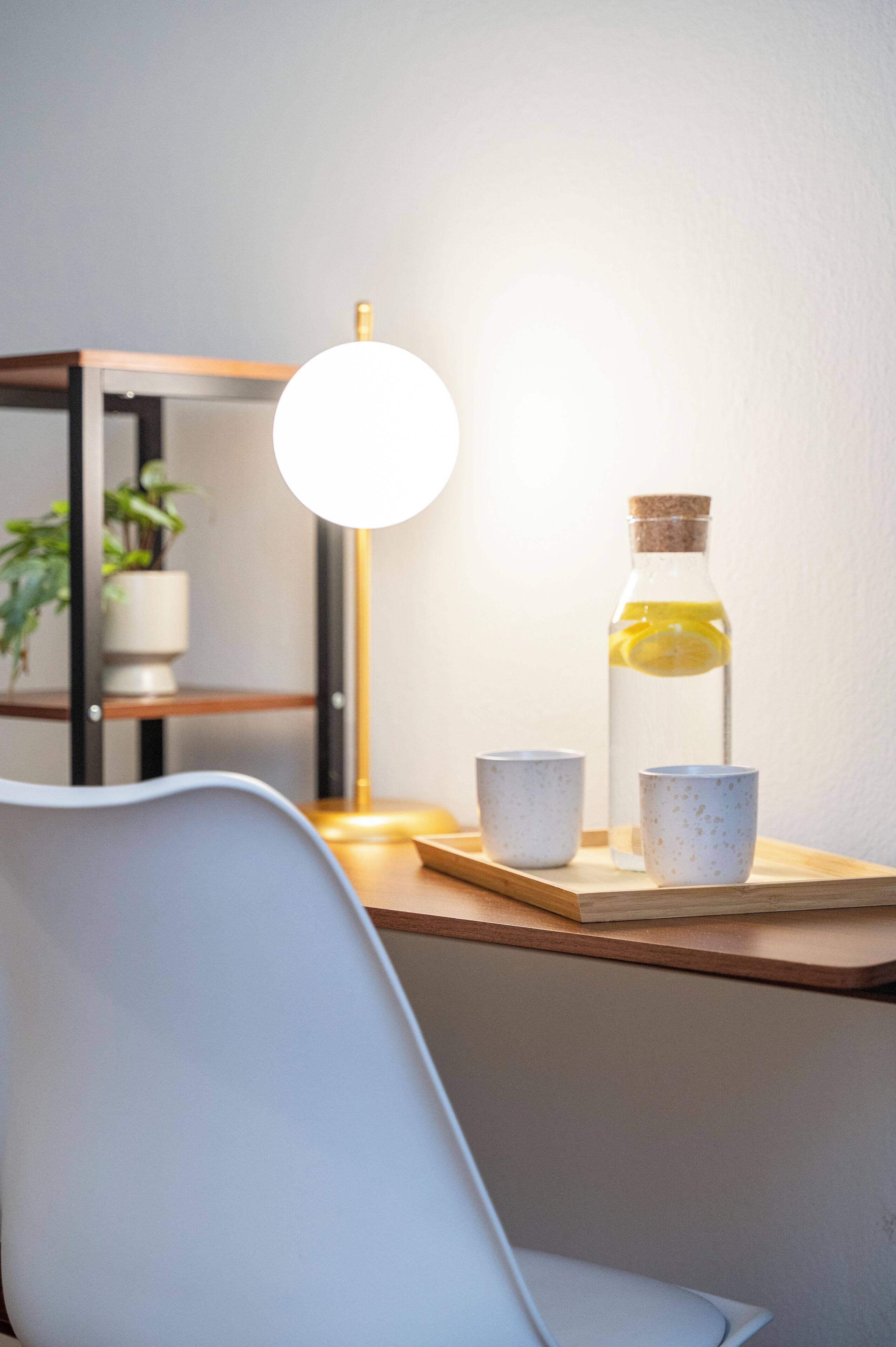 Une pièce moderne avec une chaise blanche, une table en bois, une bouteille en verre munie d’un bouchon en liège et une lampe blanche.